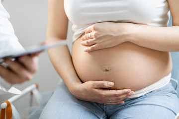 Pregnant woman hugging her belly, sitting on a gynecological chair during a medical examination, cropped view without face focused on abdomen