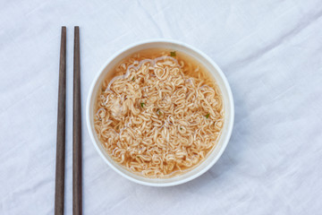 Top view of Instant noodles in a white bowl and chopsticks on a white tablecloth.