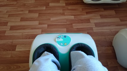 White with turquoise electric foot massager in the process of operation on a background of linoleum with imitation laminate brown. Legs in gray sweatpants are in an electric massager.