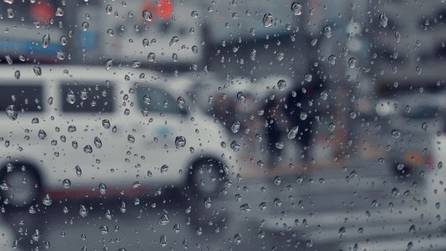 Rain flows on the side glass window of car. Blurry car and traffic on the road. Bokeh people with umbrellas waiting crossroad on the walkway. Cinema scene in slow motion