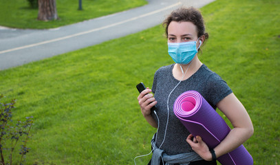 Active sport and fitness lifestyle during quarantine, coronavirus, covid-19. Young caucasian fitness woman in medical mask with exercise mat, smartphone and headphone before workout outdoor