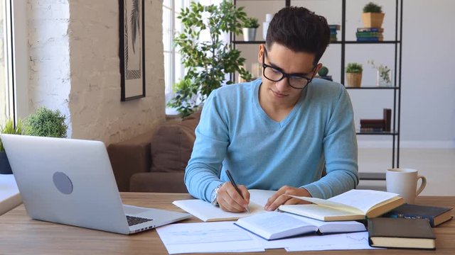 Ethnic young man in glasses writing in notebook then looking at camera and smiling while sitting at table with laptop and doing homework at home