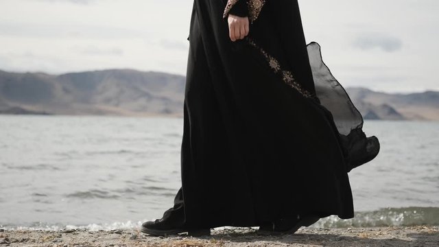 caucasian girl walks along lake shore, in black mourning dress,woman walks on rocks, near mountains