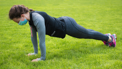 Fototapeta na wymiar Sport during quarantine, coronavirus, covid-19 concept.Young exercising fitness woman in medical protective mask doing plank or pushups on green grass in park.Caucasian girl training crossfit 