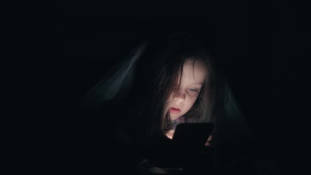 Child night phone. Late online chat. Little girl hiding under blanket texting message in darkness.