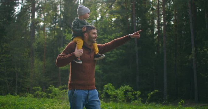 Slow motion: Son sitting on his fathers neck in the spring sunny park. Fathers day. Father and boy walk in forest and son on his neck. Father and son went on a forest adventure together.