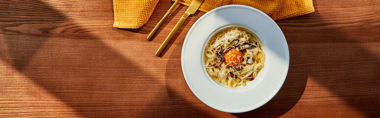 top view of delicious pasta carbonara served with golden cutlery and yellow napkin on wooden table in sunlight, panoramic shot