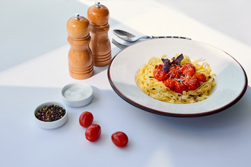 delicious pasta with tomatoes served with cutlery, salt and pepper mills on white table in sunlight