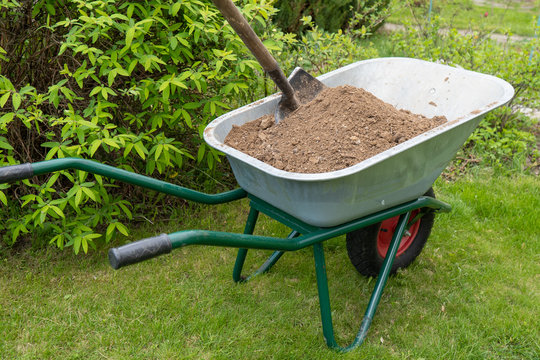 Old wheelbarrow on green grass with soil and a shovel. Back view