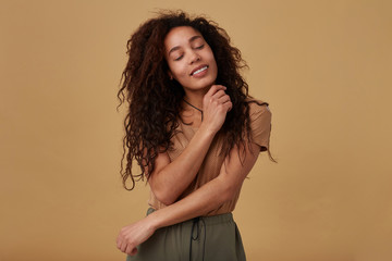 Goog looking young glad brown haired curly dark skinned woman keeping her eyes closed while smiling pleasantly and embracing her with raised hands, isolated over beige background