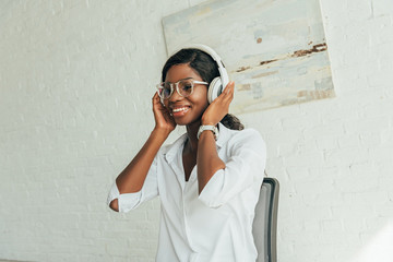 happy african american girl in eyeglasses touching wireless headphones while listening music