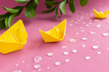 Yellow spring paper ships, boats floating in sparkling drops of water on pink,purple background.Branch of green leaves.Summer concept.Greeting card.Crafts, DIY family activity entertainment with kids
