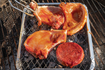 Fresh meat seen being laid on a camping BBQ grill which is placed on top of a regular garden BBQ. Tongs are seen laying chops on the grill.