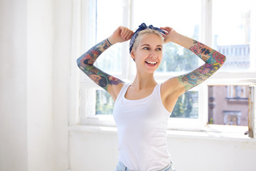 Cheerful young pretty blonde female with tattoos wearing headband and white shirt while posing in front of big window, looking aside with charming smile