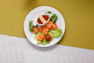 prawns on skewers, grilled. served on lettuce leaves with cherry tomatoes, lemon and hot sauce. in a white round plate set on a white wooden tabletop covered with a mustard-colored tablecloth