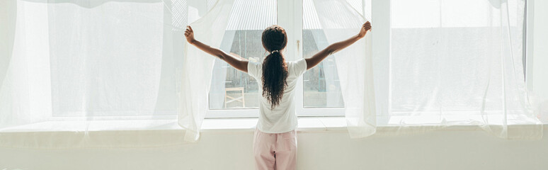 back view of african american girl in pajamas opening window curtains in sunshine, horizontal image