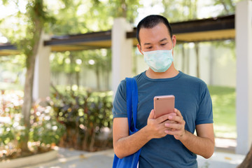 Young Asian man using phone with mask for protection from corona virus outbreak in nature outdoors