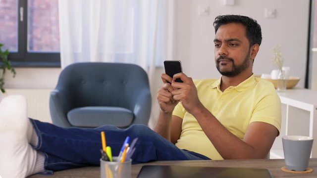 technology, remote job and people concept - indian man with smartphone resting feet on table at home office