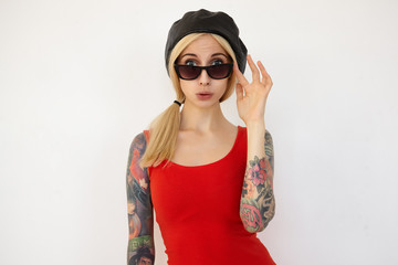 Studio shot of young attractive tattooed blonde woman with ponytail hairstyle looking surprisedly at camera while posing over white background in black beret and red dress