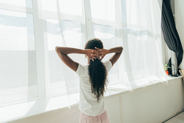 back view of african american girl standing by window in sunshine with hands behind head