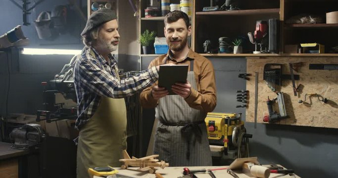 Two carpenters standing in workshop and watching some video on tablet device. Family carpentry business. Father and son handicraftsmen watch something on tablet computer in workroom.