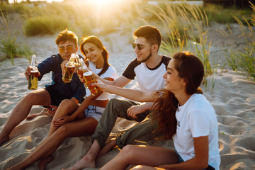 Group of young friends sitting together at the beach talking and drinking beers at sunset. Summer holidays, vacation, relax and lifestyle consept.