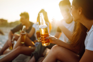 Group of young friends sitting together at the beach talking and drinking beers at sunset. Summer holidays, vacation, relax and lifestyle consept.