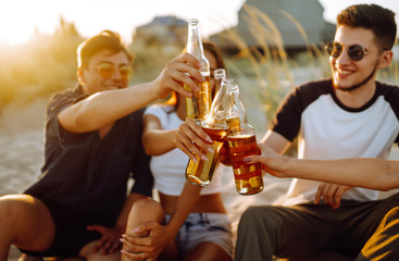Group of young friends sitting together at the beach talking and drinking beers at sunset. Summer holidays, vacation, relax and lifestyle consept.