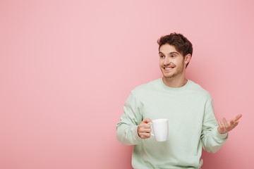 cheerful young man holding cup while standing with open arm and looking away on pink background