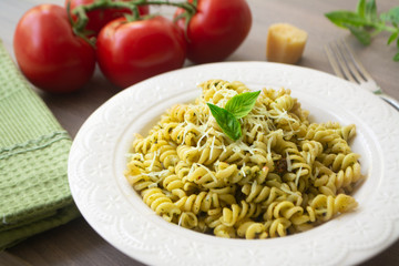 Traditional Italian vegetarian pasta fusilli with fresh homemade basil and pine nut pesto sauce and topped with grated Parmesan cheese. Tomato, basil, parmesan cheese, napkin and fork in background