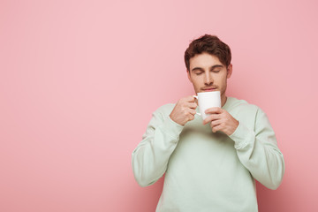 handsome young man enjoying flavor of coffee while holding white cup on pink background