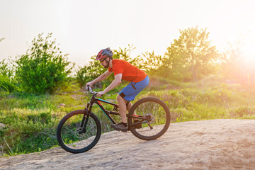 Fototapeta na wymiar A cyclist in an orange T-shirt and helmet rides a mountain bike at sunset time.