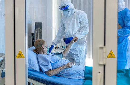 Elder Caucasian Female Patient Gets Blood Pressure Checked By Team Of Medical Doctors In Quarantine Room. Medical Staff Wear Sterile Personal Protective Equipment Suit And Face Shield. COVID-19 Check