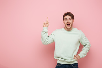 excited guy showing idea sign while standing with hand on hip and looking at camera on pink...