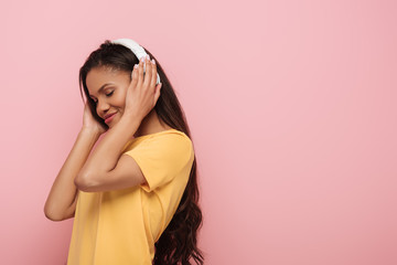 smiling african american girl listening music in wireless headphones with closed eyes isolated on pink