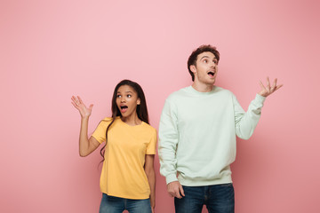 shocked interracial couple standing with open arms while looking away on pink background