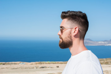 Profile portrait of a man with sunglasses, a beard and a white shirt, with the sky and the blue ocean in the background.