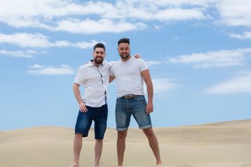Two handsome and smiling boys standing in the sand dunes with the blue sky behind.