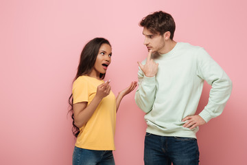 serious guy touching chin while looking at african american girlfriend quarreling on pink background