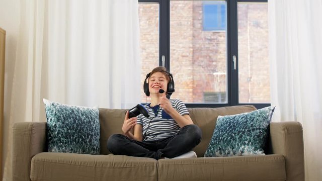 leisure, children, technology and people concept - happy boy in headphones with microphone and gamepad playing video game and celebrating success at home