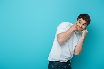 Fototapeta na wymiar displeased young man plugging ears with fingers while looking away on blue background