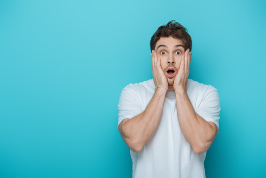 Shocked man touching face while looking at camera on blue background