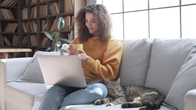 Happy young hispanic latin teen girl watching movie on laptop holding computer, laughing, drinking tea, playing cuddling with cat relaxing having fun sitting on sofa at home in cozy living room.