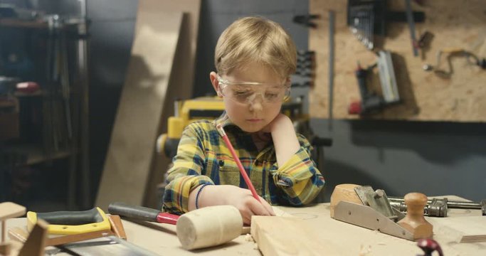 Small cute Caucasian boy in goggles working in carpentry workshop and drawing with pencil. Little schoolboy making drafts on wood while crafting with hardwood.