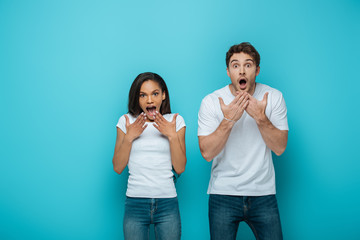 shocked interracial couple holding hands near faces with open mouths while looking at camera on blue background