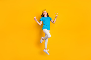 Fototapeta na wymiar Full body photo of funny little lady jumping high good mood cheerfully meet friends say hello show v-sign wear casual blue t-shirt headband trousers shoes isolated yellow color background