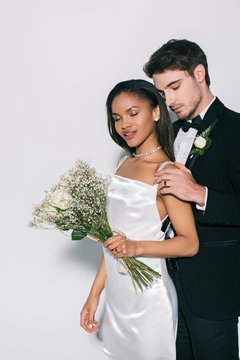 handsome groom touching shoulder of beautiful african american bride holding wedding bouquet on white background