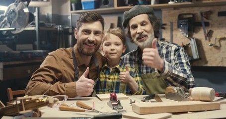 Portrait of three male generations smiling to camera and giving thumbs up. Grandfather, father and son of cheerful carpenters in workshop. Old man with son and grandson. Family of joiners.