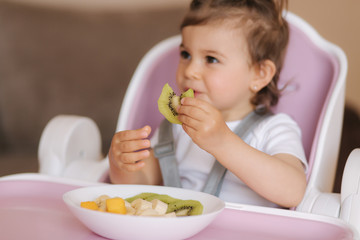 Portrait of happy little kid eating kiwi in high chair. Healthy nutrition for kids