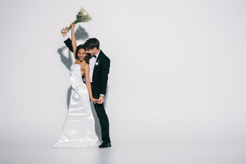 full length view of young, elegant interracial newlyweds dancing on white background